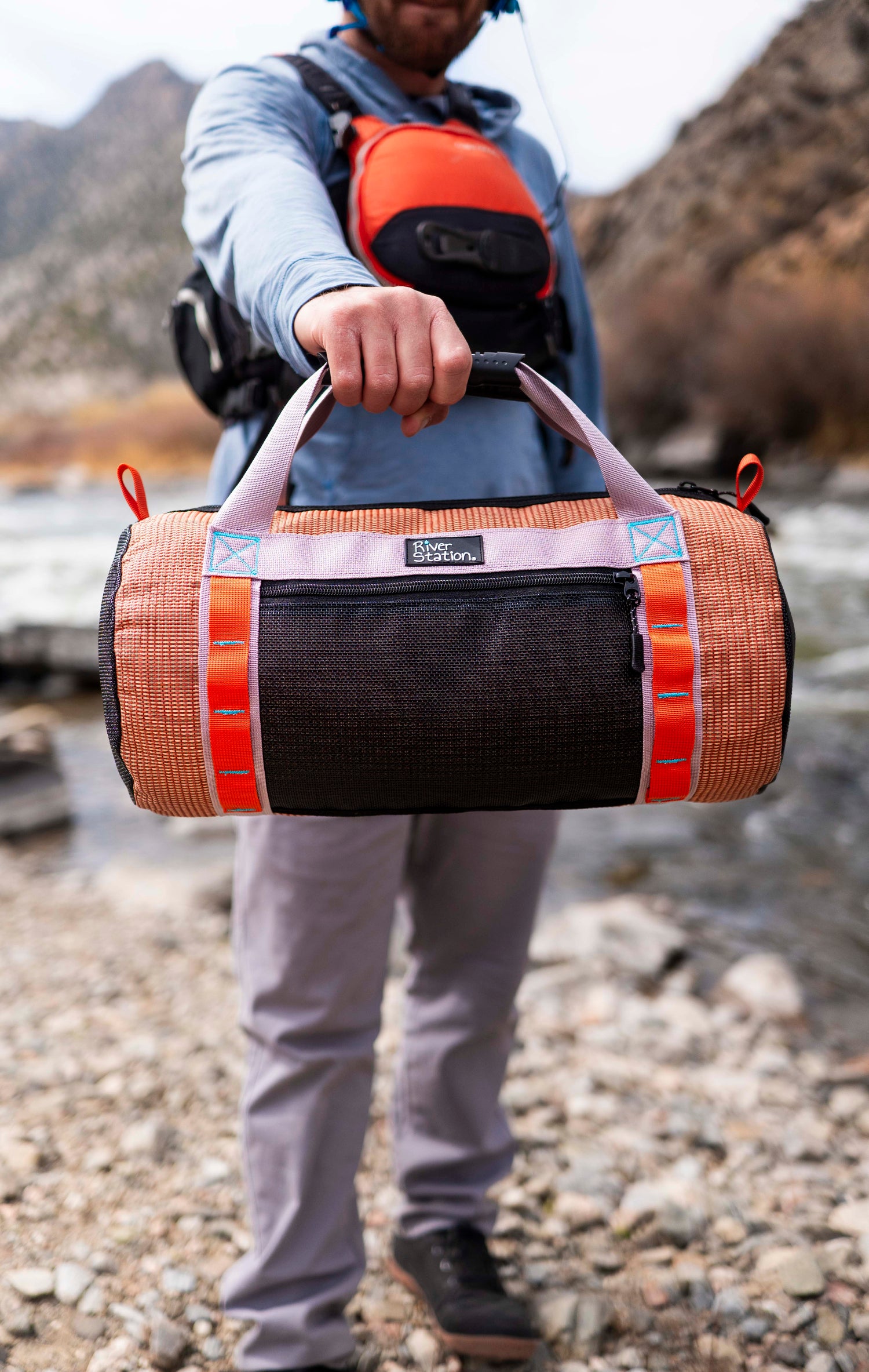 Person holding a duffel bag outdoors near a river with mountains in the background