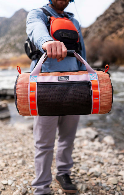 Person holding a duffel bag outdoors near a river with mountains in the background