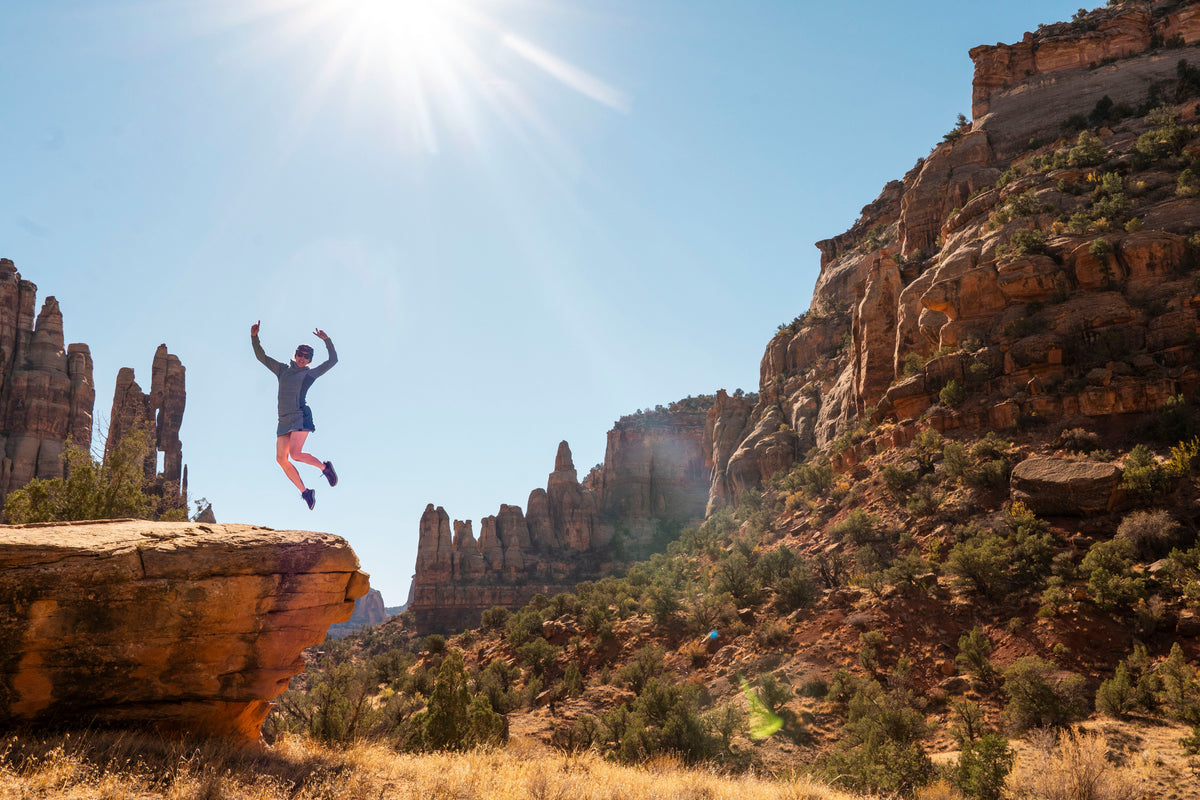 A person jumps from a rocky ledge in a desert canyon landscape under a bright sun and clear blue sky. The scene features tall orange-brown rock formations and dry grass.